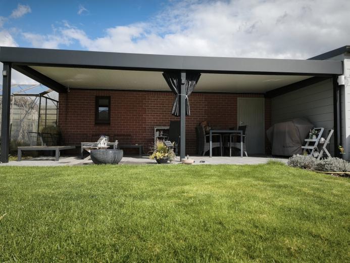 Luxe antraciet aluminium terrasoverkapping aan een bakstenen muur met moderne lounge-set en verlichting.