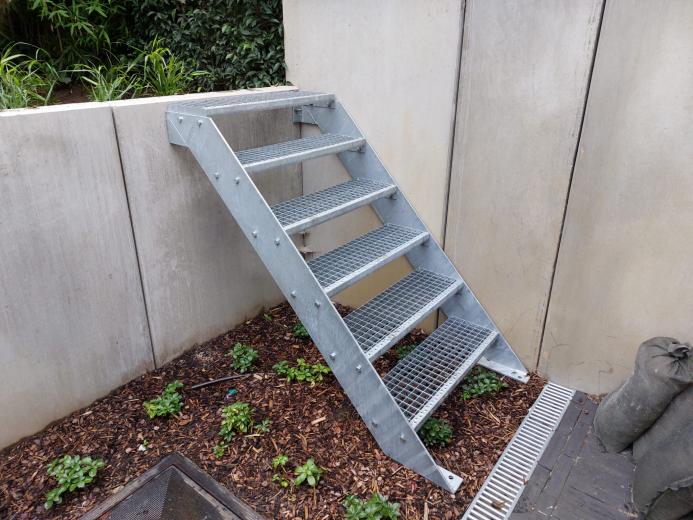 Zijaanzicht van een verzinkte stalen buitentrap met roostertreden gemonteerd tegen een betonnen keerwand.