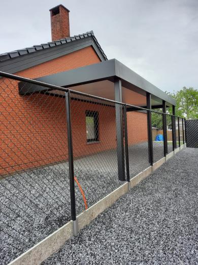 Modern antraciet aluminium carport overkapping gemonteerd aan de gevel van een bakstenen huis met grijze grind oprit en zwart gaashekwerk.