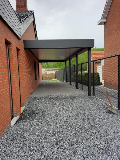 Hoekaanzicht van een moderne antraciet aluminium overkapping carport tegen een rode bakstenen gevel met grijze dakpannen, met zwart gaashekwerk op een betonnen rand en grind op de voorgrond.