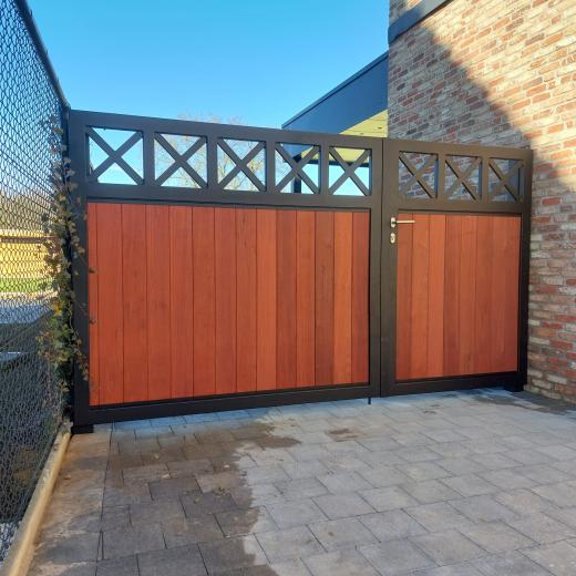 Zwarte metalen draaipoort met verticale houten planken en decoratieve kruisen aan de bovenzijde, gemonteerd naast een bakstenen muur.