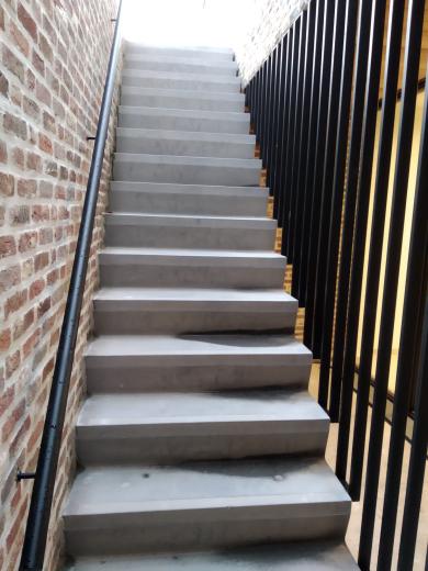 Zijaanzicht van een moderne zwarte stalen trapleuning gemonteerd op een bakstenen muur, naast een betontrap met een spijlenbalustrade.