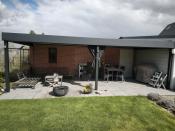 Luxe antraciet aluminium terrasoverkapping aan een bakstenen muur met moderne lounge-set en verlichting.