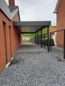 Hoekaanzicht van een moderne antraciet aluminium overkapping carport tegen een rode bakstenen gevel met grijze dakpannen, met zwart gaashekwerk op een betonnen rand en grind op de voorgrond.