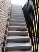 Zijaanzicht van een moderne zwarte stalen trapleuning gemonteerd op een bakstenen muur, naast een betontrap met een spijlenbalustrade.