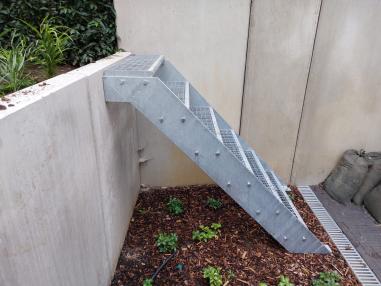 Zijaanzicht van een verzinkte stalen buitentrap met roostertreden gemonteerd tegen een betonnen keerwand.