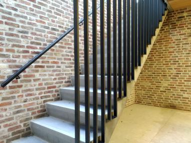 Zijaanzicht van een moderne zwarte stalen trapleuning gemonteerd op een bakstenen muur, naast een betontrap met een spijlenbalustrade.